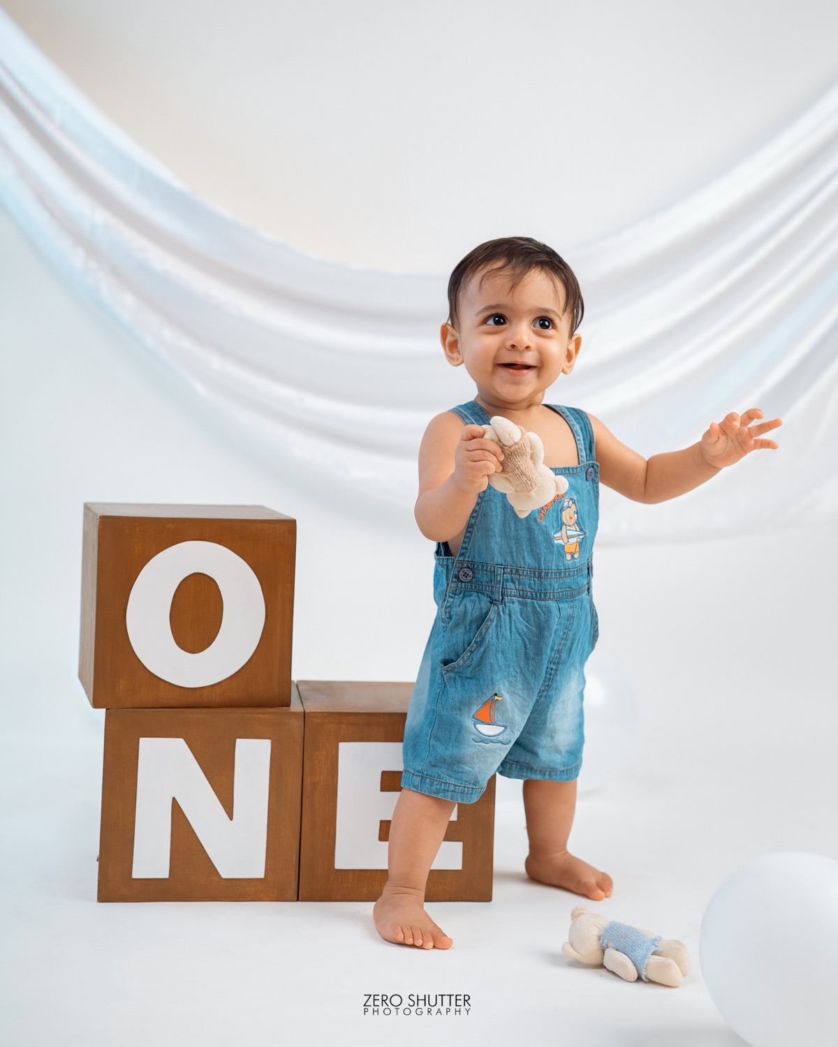 Smiling one year old in denim dungarees beside wooden ONE blocks