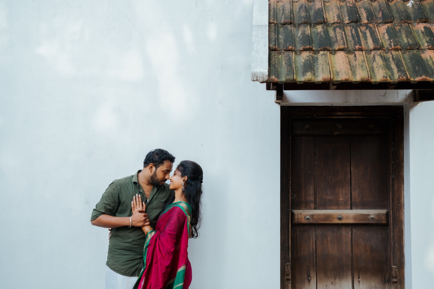 Couple sharing a nose-to-nose moment beside a white wall and old wooden door