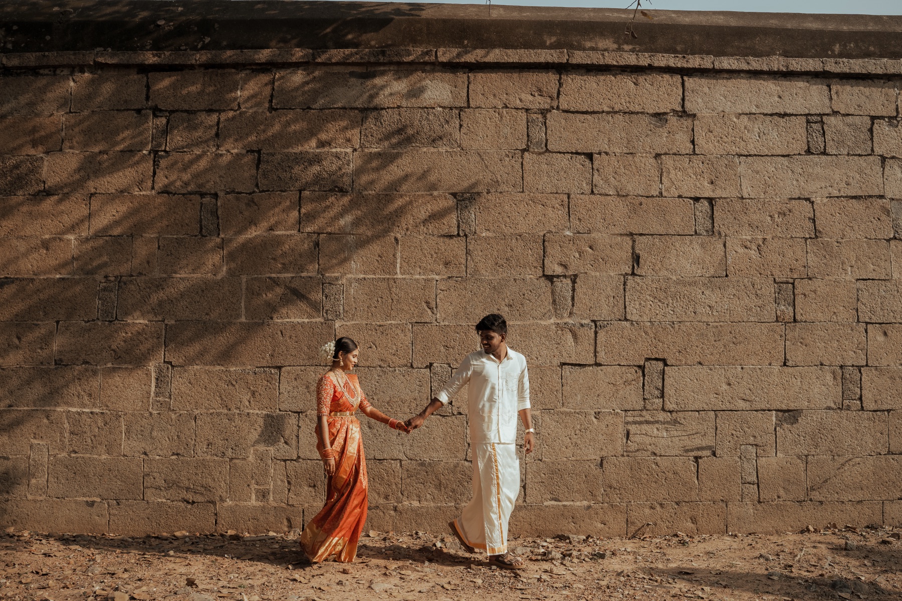Bride and groom walking hand in hand along an old stone wall